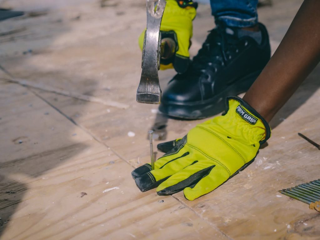 A worker wearing bright gloves uses a hammer on a plywood surface, emphasizing safety and construction.